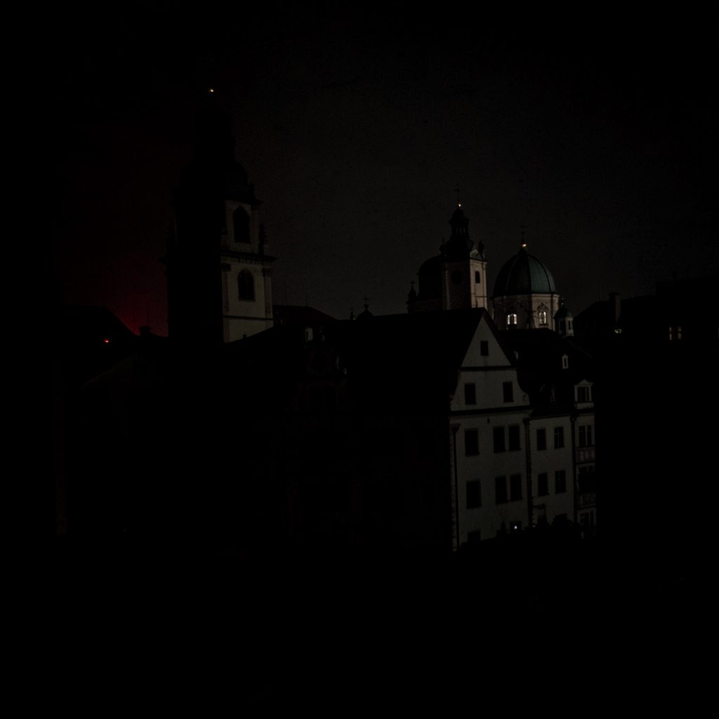 Dark, low-key emblem-style graphic in deep red and black tones evoking a civic coat of arms, moody and shadowed